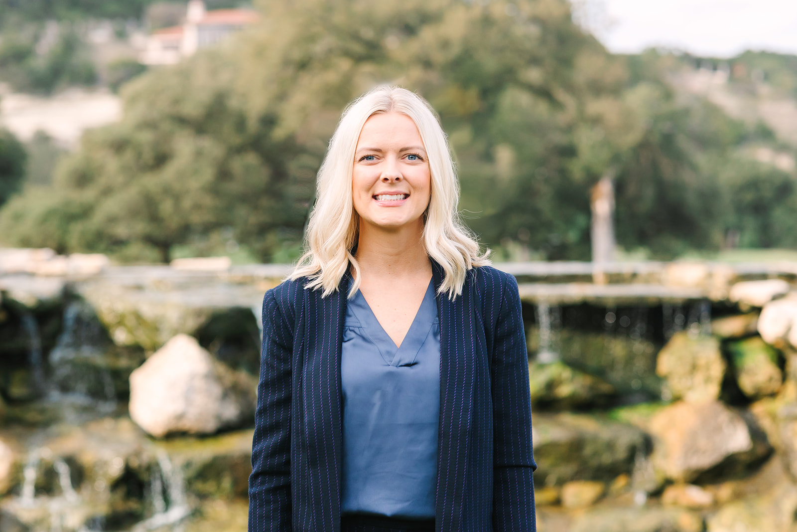 A blonde woman in a business suit standing in front of a waterfall.