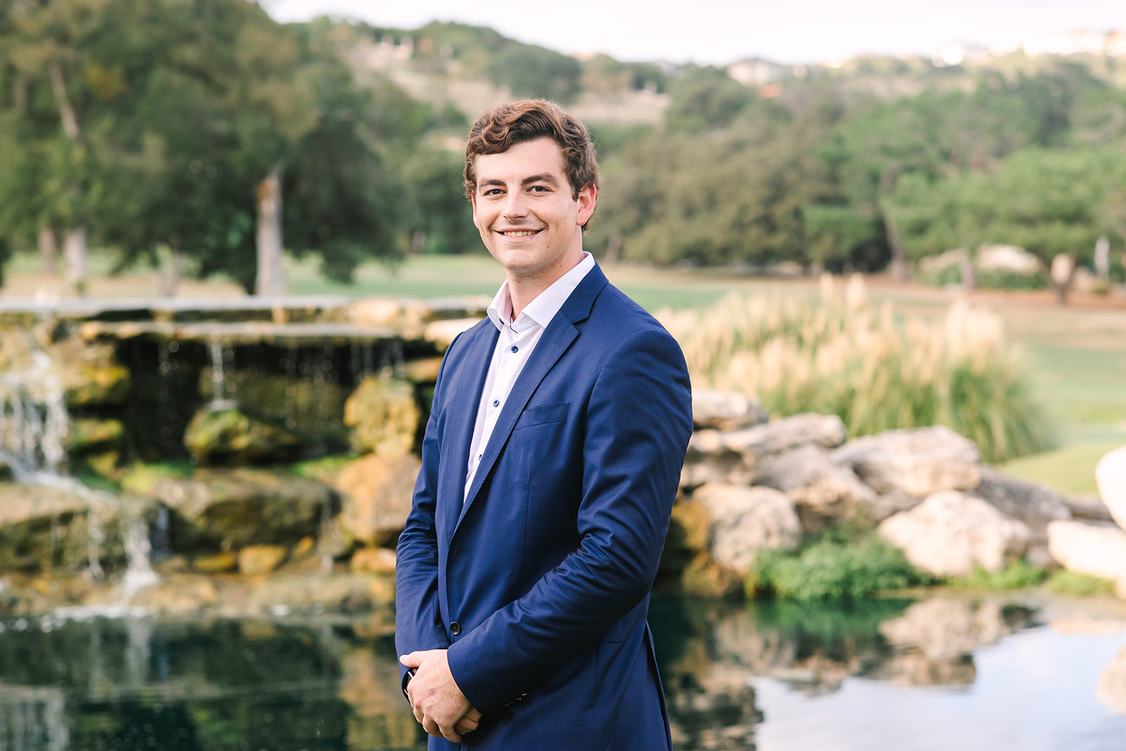 A young man in a blue suit standing in front of a pond.